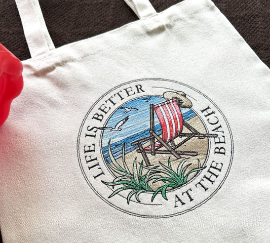 White tote bag with beach-themed embroidered design and text 'Life is better at the beach' on a 100% cotton tote bag.