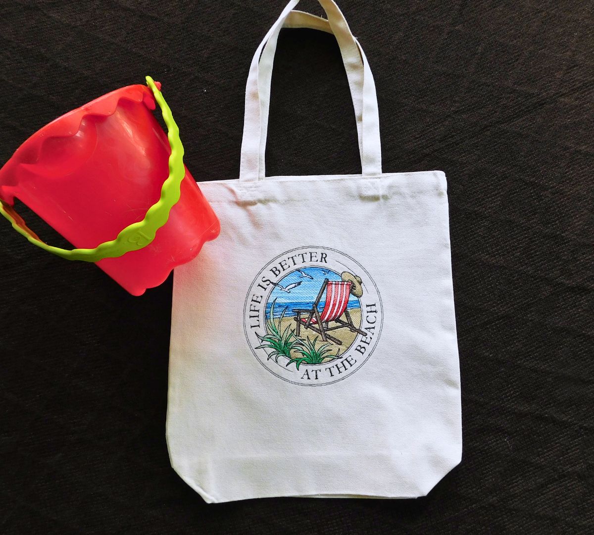 White embroidered tote bag with beach-themed embroidery design and red sand bucket on a dark background