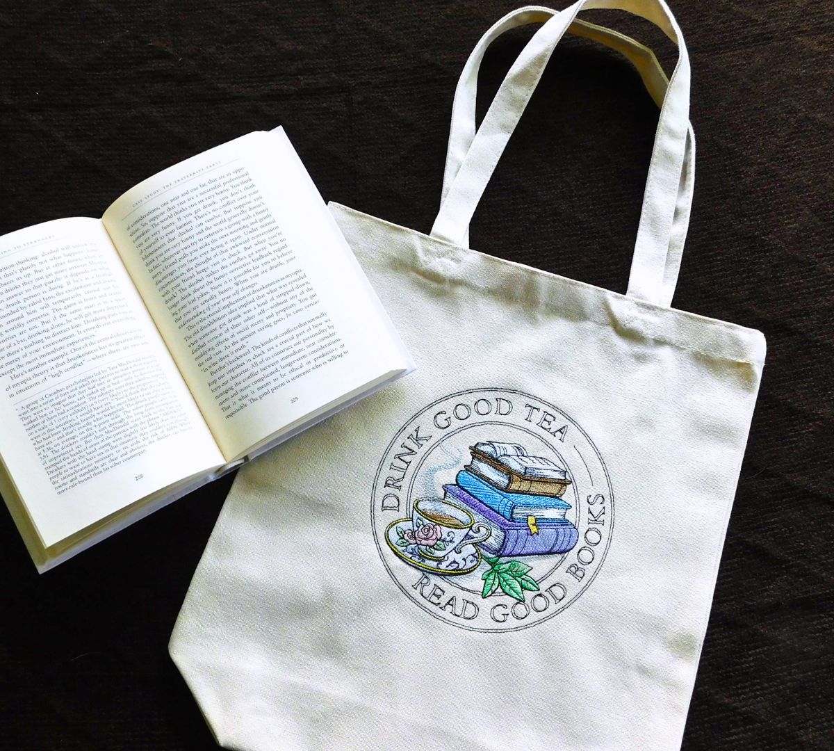 White embroidered tote bag with 'Drink Good Tea, Read Good Books' design next to an open book on a dark surface