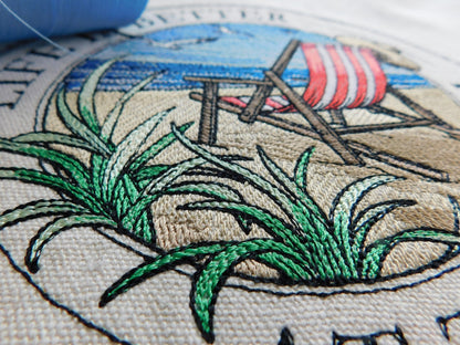 Embroidery of a beach scene with grass, lounge chairs, and an red striped beach chair tote bag.