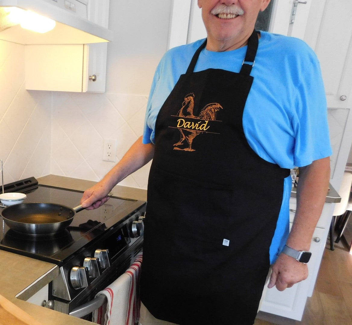 Man in a kitchen wearing a personalized embroidered black apron with 'David' printed on it, holding a frying pan.