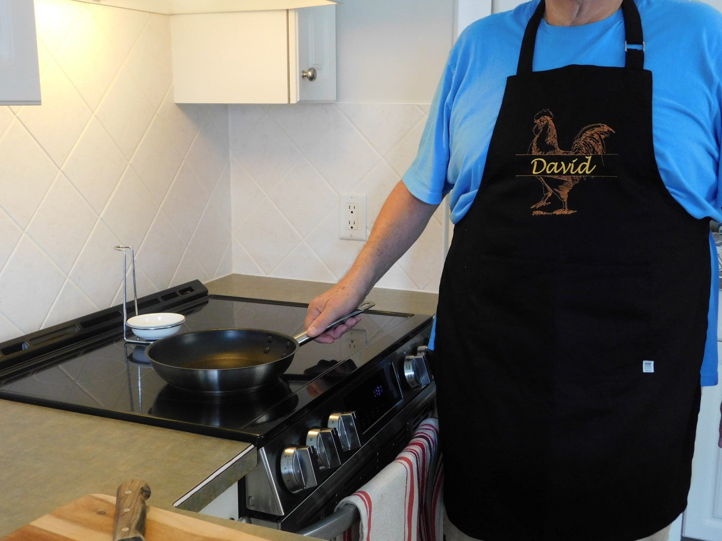 Person wearing a personalized embroidered black apron with 'David' printed on it, holding a frying pan.