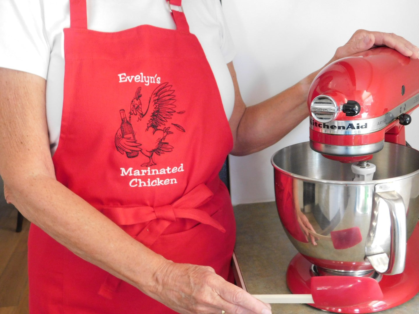 Person wearing a personalized embroidered red apron with a chicken graphic using a red KitchenAid mixer.