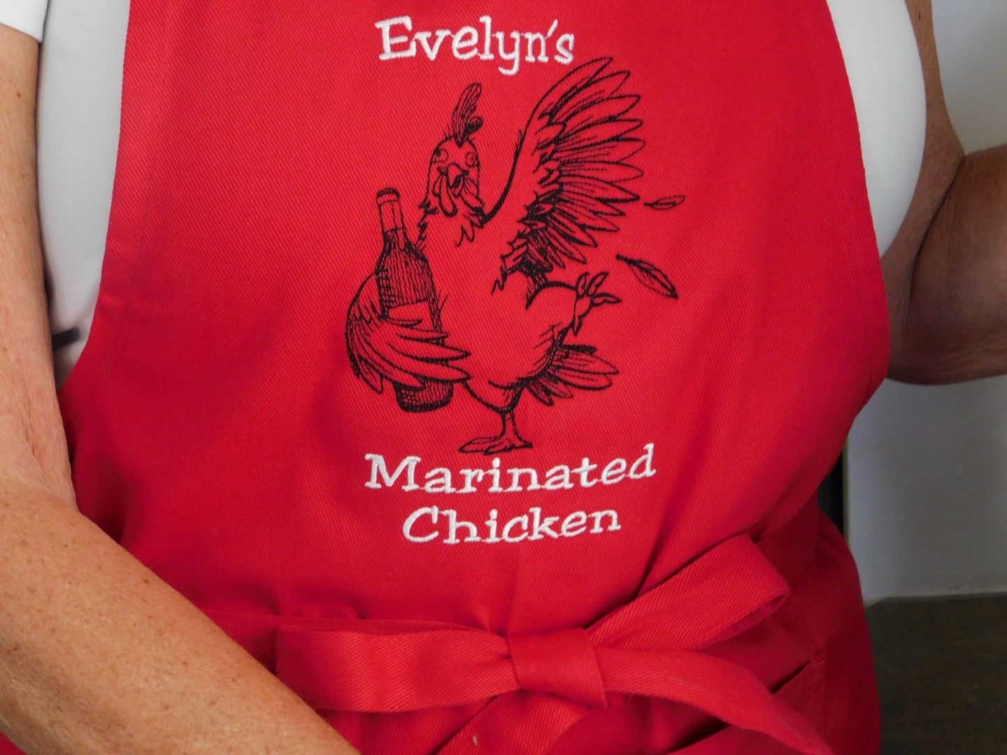 Personalized embroidered red apron with a graphic of a rooster and text 'Evelyn's Marinated Chicken' on a person wearing it.